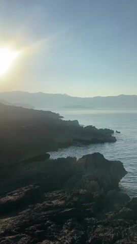 The video displays a scenic coastal view at sunrise. The sun is positioned in the upper left corner, casting a warm glow. In the foreground, there are dark, jagged rocks lining the shore. The sea is calm and stretches out towards a distant, hazy shoreline with mountains in the background. The sky is clear and a soft blue. There are no people or explicit actions depicted in the video. The overall ambiance is peaceful and serene.