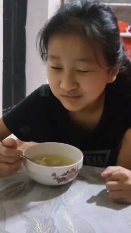 A young girl with dark hair pulled into a ponytail, wearing a black t-shirt, sits at a table with a white tablecloth. She is holding a spoon in her right hand, which is submerged in a white bowl of clear broth containing what appear to be dumplings or noodles. The girl is looking down at the bowl and appears to be eating the soup. Her left hand rests on the table. The background is dimly lit, with a portion of a white wall and a dark doorway visible.