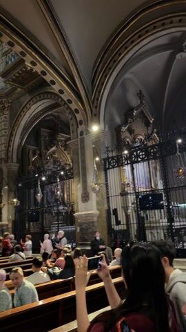 The camera captures the interior of a grand church, teeming with visitors seated on pews and standing around. The church's architecture is characterized by high, arched ceilings, ornate decorations, and a central dome. The walls are adorned with intricate patterns and designs. The camera moves to provide a view of the entire church. Most of the people are dressed casually, and are standing or sitting looking at the interior of the church. Some of them are taking photographs.
