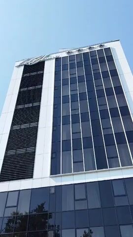 The video shows a low-angle view of a modern hotel building with a white facade and a large glass-covered lower section. The hotel's name, "Le Blanc HOTEL," is visible above the entrance, along with what appear to be star ratings. The building has a dark, reflective glass facade on the lower floors, and a section with horizontal louvers on the upper floors. Two flags, one with stars and stripes and another with horizontal stripes, are mounted on poles near the entrance. The sky is clear and blue. The video is shot in portrait orientation and appears to be a static shot of the hotel exterior.