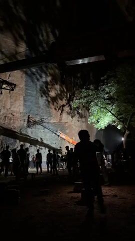 The video shows a group of people gathered at the base of a large, rocky cliff face at night. A large orange crane with a long boom is positioned in front of the cliff. The boom of the crane extends up the cliff face, and at its end is a large, metallic structure that appears to be a camera rig or some form of stage equipment. Several figures, silhouetted against the dim lighting, are visible standing around the base of the cliff and near the crane. Lights illuminate parts of the cliff face and the surrounding area, creating stark shadows. The ground appears to be uneven and dusty. The overall impression is of a film or event production set up in a rugged outdoor environment at night.