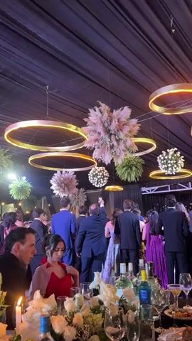A low-angle shot shows a wedding reception from a table looking towards a dance floor. The ceiling is draped with black fabric and illuminated by circular gold lights with orange inner halos. Floral arrangements, including large fluffy pampas grass and smaller white floral spheres, hang from the ceiling. In the background, a group of guests in formal attire, many in suits and dresses, are gathered. Some are dancing or socializing. A woman in a red dress sits at the table in the foreground, looking down. The table is set with plates, glasses, wine bottles, and a large floral centerpiece.