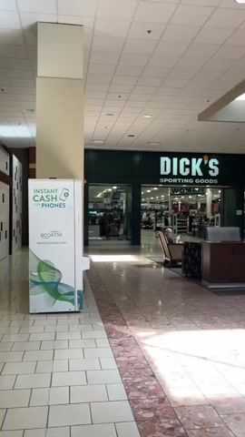 The video shows a view of a shopping mall interior. On the left, there is a white ATM machine with green graphics and the words "INSTANT CASH FOR PHONES" and "ECOATM" visible. To the right of the ATM is a large white column. Further to the right, a store with a green sign reading "DICK'S SPORTING GOODS" is visible. In front of the store, there is a seating area with chairs and a low dark brown counter. A person is seated in a chair on the left side of the seating area, looking at something in their lap. Another person is standing to the right of the seated individual. The floor is tiled with a pinkish marble pattern. The ceiling is white with square light fixtures. The overall lighting suggests daytime.