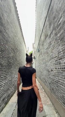 A person walks down a narrow alleyway between two tall brick buildings. The person is wearing a black top and black pants. The alleyway has a stone path. The person walks away from the camera, then turns to face the camera and walks towards it, then turns back around and walks away again.