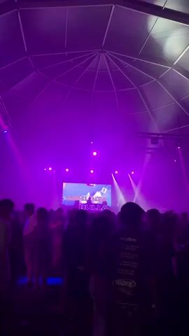 A wide shot captures an indoor music event with a large crowd of people dancing. The ceiling is a geodesic dome structure with a spotlight illuminating the space. A stage with a large screen displaying text and graphics is visible in the background, accompanied by flashing purple and green lights. The crowd is diverse, with individuals of various ages and ethnicities. The atmosphere is energetic and lively, with people moving and interacting with each other. The lighting is dim, with the primary illumination coming from the stage and spotlights.