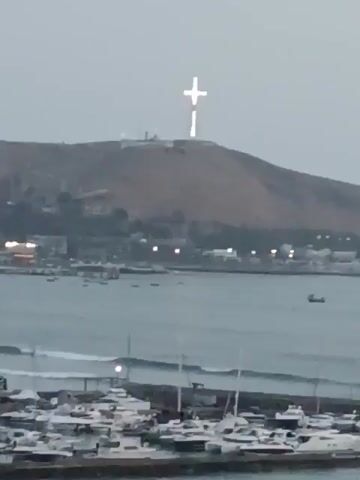 The video shows a wide shot of a harbor with a large hill in the background. On top of the hill stands a large, illuminated cross. In the foreground, a marina is filled with boats, mostly white, bobbing gently in the water. The water in the harbor appears calm with small waves. In the middle ground, a coastal city is visible, with buildings lit up with lights. The sky is overcast, casting a soft light over the scene. The camera is static.
