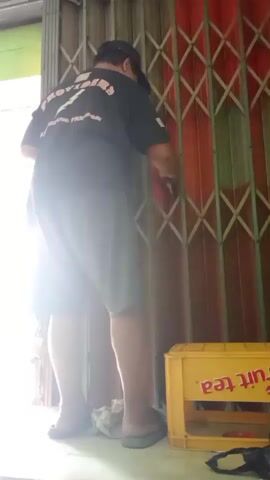 A person wearing a black t-shirt, grey shorts, and a black cap is standing in front of a metal accordion gate. The person is holding a white object and appears to be cleaning or wiping the gate. To the right of the gate, there is a yellow crate with the words "fruit tea" visible. The environment appears to be outdoors, possibly near a building entrance, with bright light coming from the left.