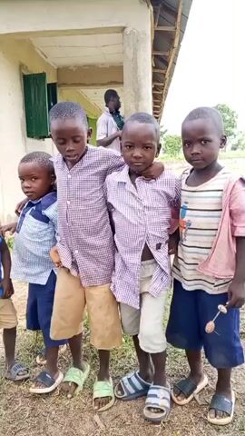 Four young boys stand in a line in front of a building. The building appears to be a schoolhouse with a light-colored wall and a corrugated metal roof. Two of the boys are wearing plaid shirts and shorts, one is wearing a collared shirt with a blue and white pattern and shorts, and the other is wearing a striped t-shirt and shorts. They are all wearing sandals. In the background, a young boy is standing near the building. The ground is covered in dirt and some dry grass.