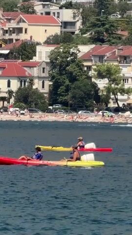 The video shows three kayaks on a body of water. The closest kayak is red and yellow, with two people in it. The person in the front is wearing a blue shirt and a hat and is paddling. The person in the back is wearing a dark shirt and a hat and is also paddling. Behind them is a yellow kayak with one person in it, wearing a dark shirt and hat and paddling. Further behind is a red kayak with one person in it. In the background, there is a beach with many people sunbathing, cars parked, and buildings on a hill. The water is blue and appears to be choppy.