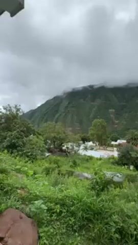 The video shows a view of a lush green mountain under a cloudy sky. The mountain is covered with vegetation and has some visible layers of red soil. The top of the mountain is partially obscured by clouds. In the foreground, there are trees and bushes. There are also some buildings visible in the distance. The overall impression is of a natural landscape with a somewhat overcast atmosphere.