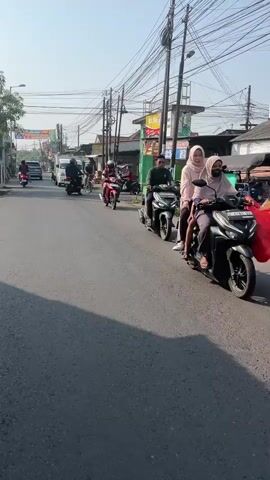 A person is riding a red motorcycle down a paved road. The person is wearing a white helmet and a dark jacket. There are other motorcycles and vehicles on the road, as well as buildings and power lines. The sky is clear and the sun is shining.