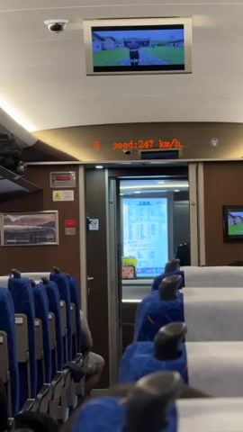 The video shows the interior of a train car and the view from its window. The car has blue seats with an orange stripe and a headrest cover. In the first shot, a screen overhead displays a video, and an illuminated sign shows the train's speed as 247 km/h. The door leading to the next car is visible, as well as various posters and information panels. The second part of the video shows the view from the train window. The scenery includes buildings, trees, and hills, with the view slightly blurred due to the speed of the train.