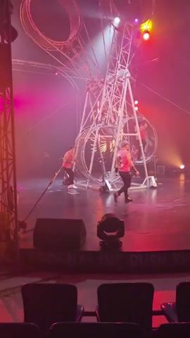 The video captures a thrilling performance on a stage, likely in Kathu, Thailand. The main focus is a large, complex metal structure resembling a giant Ferris wheel with multiple rotating cages or wheels. Several performers, dressed in bright pink tops and dark bottoms, are actively engaged in a dynamic act within these spinning structures. Some are inside the rotating wheels, appearing to run or balance, while others might be manipulating the apparatus. The stage is bathed in dramatic lighting, with a heavy use of pink and red hues, creating a smoky and atmospheric effect. Yellow spotlights cut through the haze, illuminating the performers and the intricate machinery.

In the foreground, there are large black speakers and stage lighting equipment, suggesting a live performance setting. The audience's view is partially obstructed by what appears to be the edge of a seating area, indicating the video was taken from the audience. The overall mood is one of excitement and high energy, characteristic of a circus or theatrical show. The specific time of day is not discernible, but the indoor setting and stage lighting point towards an evening performance. There is no visible weather to comment on. While the location is contextually given as Kathu, Thailand, no explicit text or landmarks within the video confirm this.