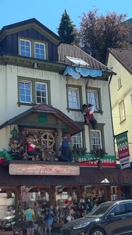 A playful animated figure of a bear wearing a hat, red vest, and black pants is suspended by wires on the side of a light green building. The bear is holding a camera and appears to be taking a picture. The building has multiple windows, some decorated with white trim and green shutters. Above the bear, a decorative element resembling clouds with strings of lights hangs from the roofline. Below the bear, on a lower level of the building, two other animated bear figures are visible. One bear stands and wears a blue vest and dark skirt, while another bear sits and appears to be working with gears and machinery. A sign on the building reads "Uhren 1000 Clocks Souvenirs." The scene is outdoors on a sunny day, with trees visible in the background. A black car is parked in the foreground, and other people are walking on the sidewalk.