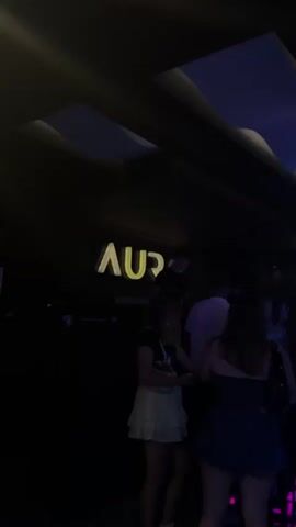 The video shows a dimly lit interior that appears to be a nightclub or bar. People are visible in the background, some standing and others seemingly dancing. The lighting is primarily composed of purples and blues, with some white accent lights on the ceiling. A sign with illuminated letters that spell "AURA" is visible on a dark wall. There are tables and chairs scattered throughout the space. The camera is positioned at a low angle, looking upwards towards the ceiling and the sign. The overall atmosphere suggests a lively social gathering.