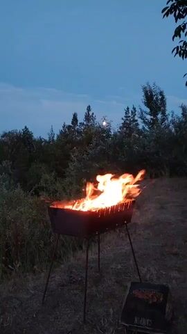 A fire burns in a metal barbecue grill, which is on a tripod stand. The grill is outdoors, with trees and foliage in the background. The sky is dark, indicating it is either dusk or night, and there is a full moon visible in the sky. A bag of charcoal is on the ground next to the grill. The fire is large and vibrant, with flames reaching upwards. The scene appears to be a campsite or a backyard gathering.