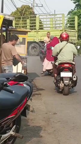 The video shows a street scene, likely in Indonesia, with several motorbikes and food vendors. In the foreground, a black and red motorbike is parked. To the left, a food stall with a yellow banner advertising "SATE AYAM KAMBING RUL Madura" is visible, with a vendor attending to it. Several people are on motorbikes, some carrying passengers. One motorbike in the middle ground has a woman wearing a green shirt and blue jeans, with a child sitting in front of her. Another motorbike further back carries a man, a woman, and a child. A green truck is seen in the background. The scene appears to be a bustling street with food stalls and traffic.