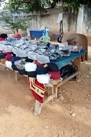 A street vendor is selling a variety of taqiyahs, or skull caps, on a wooden table outside. Some are plain in color, and others are decorated with embroidery and are wrapped in plastic. A man in a tan outfit is standing in the background next to the table. The ground is dirt, and there is a cinderblock wall in the background.