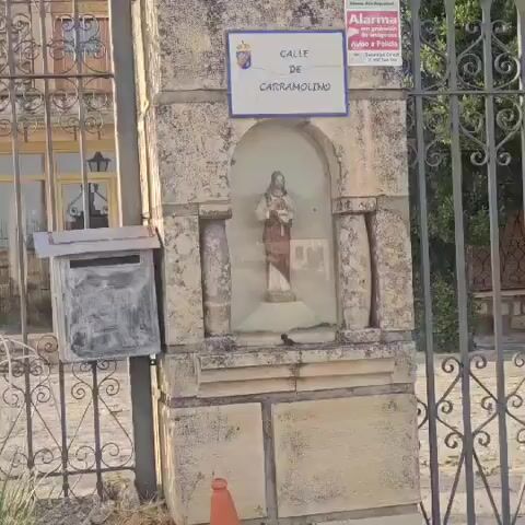 A video shows a weathered stone pillar with a mailbox and a small shrine. Above the shrine is a sign that reads "CALLE DE CARRAMOLINO". Behind the pillar is a black metal fence. A house is visible behind the fence, with a dark roof and windows. To the left of the pillar is a tall, multi-colored wind sculpture. The ground is covered in dry grass and weeds. The sky is clear and blue. The camera pans left to right, showing more of the fence and the house. There are no people visible in the video.