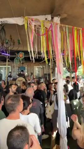 The video shows a large crowd of people gathered under an open-air structure decorated with colorful streamers. A band is performing on a raised platform at one end of the space. People are dancing and moving to the music, and the atmosphere appears lively. The structure has a rustic feel, with visible support beams and a partially covered roof. There are a variety of people in the crowd, of different ages and clothing styles.