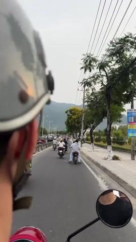 The video shows a first-person perspective from a person riding a motor scooter, likely in Vietnam. The rider is wearing a gray helmet and traveling on a road alongside other motor scooters. The traffic light is green. Trees line the sidewalk on the right of the road. In the distance, there are mountains. Power lines run along the right side of the road. A sign is visible on the right side.