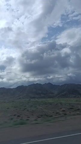 The video shows a view from a vehicle looking towards a mountain range under a cloudy sky. The foreground consists of a road and sparse vegetation. The mountains are dark and rugged, with some lighter patches of vegetation or rock. The sky is filled with a mix of dark and light gray clouds, with patches of blue sky visible. The sun is intermittently visible through the clouds, creating varying light conditions. A white and black pole is visible on the side of the road in the middle ground. The overall impression is of a somewhat dramatic, possibly stormy, natural landscape.