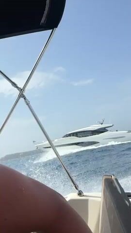 The video shows a view from a boat on a sunny day. In the foreground, parts of a boat's interior are visible, including seats and metal supports for a canopy. In the background, a large white yacht is seen moving through blue water, creating a wake. There are a few people visible on the boat from which the video is being filmed. One person, wearing a white top, is seen from the side with their arm raised. Another person is seen from the back, wearing sunglasses and holding a phone. There is also a brief glimpse of a third person's shoulder and arm. The sky is blue with some white clouds.