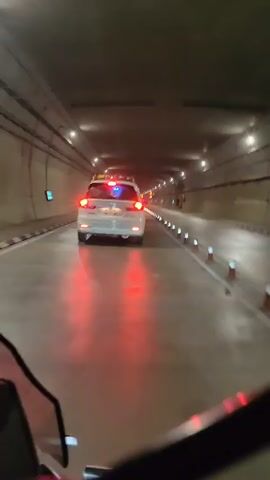 This video shows the interior of a tunnel. A white car is in front of the camera, its brake lights illuminated. The tunnel walls are dark, and the ceiling is lined with lights. Traffic cones and reflectors are visible along the side of the road. The camera appears to be mounted on a motorcycle.