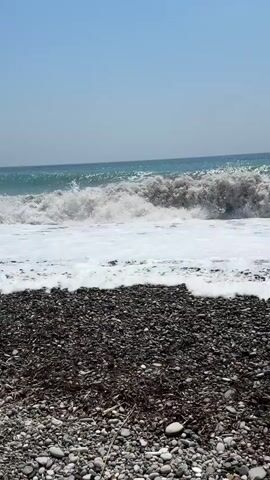 A choppy sea breaks on a pebble beach under a clear blue sky. Waves crash and foam onto the shore, with the tide reaching the edge of the beach. The water is a murky green-blue, and the beach is covered in dark grey and brown pebbles.