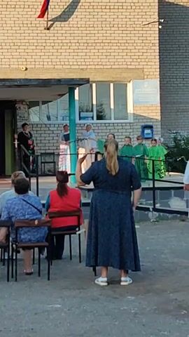 The video captures an outdoor gathering in front of a brick building, possibly a school or community center. The facade of the building features a light-colored horizontal band above the entrance, with a blue sign to the right. A Russian flag is visible at the top left of the building.

In the foreground, three women are seated on chairs facing away from the camera, observing an event taking place on a raised platform or porch. A fourth woman stands closer to the camera, facing the platform, her back to the viewer. She is wearing a dark blue patterned dress and appears to be gesturing or performing.

On the platform, several individuals are present. A woman in a floral dress stands to the left, speaking to a woman in a white outfit. Further to the right, a group of children or young people, dressed in green outfits, are standing in a line. One of them appears to be holding a microphone. An adult in white is partially visible on the far right edge of the platform.

The environment suggests a daytime event, with sunlight illuminating the scene. The ground in front of the building is paved with asphalt or concrete. The overall atmosphere seems to be one of a performance or ceremony.