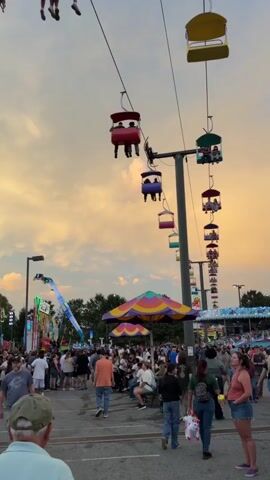 The video shows a busy amusement park or fairground at dusk. The sky is filled with a dramatic, orange and yellow sunset, with fluffy clouds. In the foreground, there is a crowd of people milling about. To the left, a tall amusement ride, painted in bright colors, is in motion, with a section resembling a barn tilting precariously. Above, a series of gondolas, part of a sky-ride attraction, are suspended from cables, carrying passengers across the scene. The gondolas are brightly colored in red, yellow, and blue. Another amusement ride, with flashing lights and a swinging arm, is visible in the background to the left. The overall atmosphere is lively and festive, with the sounds of a fair likely present.