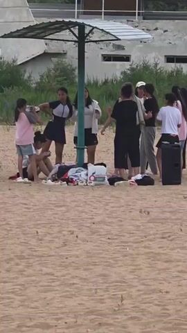 The video shows a group of people on a sandy beach, possibly near a body of water given the sandy terrain and the presence of beach umbrellas. The scene appears to be outdoors on a sunny day. The group consists of several young women and girls, dressed in casual attire suitable for a beach outing. Some are standing, while others are sitting or lounging on the sand. A large, dark speaker is visible to the right of the group, suggesting music might be playing or is intended to be played. Beach gear, including towels and bags, are scattered around the base of a large umbrella, indicating they are enjoying a leisurely day.

The people are clustered together, some interacting with each other, possibly engaged in conversation or a game. Their body language suggests a relaxed and social atmosphere. The background shows a building structure with a sloped roof and windows, partially obscured by green foliage and trees, suggesting an established area or facility near the beach. The overall mood seems to be one of leisure and enjoyment. There is no discernible text visible in the video. The time of day appears to be daytime, with the lighting suggesting it's not early morning or late evening. The setting appears to be a popular spot for recreation.