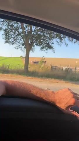 The video is filmed from inside a vehicle looking out the passenger side window. A person's arm and hand are resting on the interior of the car door, with sunlight illuminating the arm. Outside the window, a rural landscape is visible under a clear blue sky. There is a dry, grassy field with several wooden fence posts. A lone tree stands in the middle ground. In the distance, a large white pile, possibly hay or compost, is visible in the field. A power line tower is also visible on the left side of the frame. The video appears to be a casual observation of the passing scenery from a car.