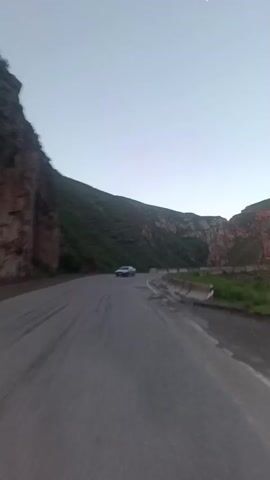 The video captures a view from a moving vehicle on a paved road winding through a mountainous area. The sky is a pale blue, suggesting either dawn or dusk, with a faint crescent moon visible. On the left side of the road, a steep, rocky cliff face rises, covered in patches of green vegetation. On the right side, the road is bordered by a concrete barrier, beyond which is a grassy slope. The road itself appears somewhat worn, with visible tire marks. In the early part of the video, a silver sedan is seen driving in the opposite direction on the road. The car then moves out of frame as the viewpoint continues along the curve of the road, the mountainous landscape dominating the scenery.