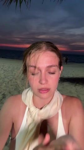 A young woman stands on a beach at dusk. The sky displays hues of pink and orange, reflecting on the sea in the background. She's wearing a white top and a light-colored scarf tied around her neck. Her hair appears wet, and she is repeatedly brushing it back from her face with her hands. Her expression is neutral as she looks directly at the camera. In the distance, other people are visible on the beach, near the water. Some palm fronds hang into the frame at the top. A small tattoo is visible on her inner left wrist.