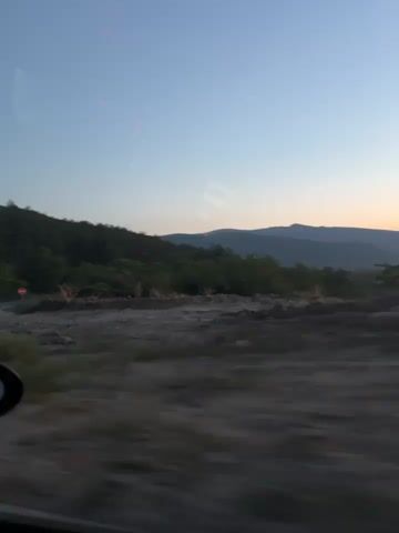This video appears to be a first-person perspective video taken from inside a moving vehicle. The scene shows a landscape during what appears to be dusk or dawn, with a clear sky fading into orange and pink hues near the horizon. There are hills and trees in the background, and the foreground is a blur of motion, suggesting the vehicle is in transit. A car's side mirror is visible in the lower left corner of the frame. The motion of the vehicle causes the scenery to blur, creating a sense of movement.