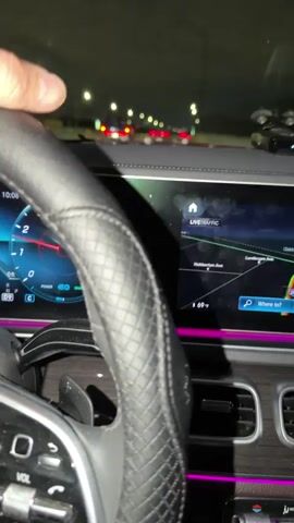 A person is driving a Mercedes-Benz at night. The dashboard displays the car's speed, which is around 70 mph, and the outside temperature is 65 degrees Fahrenheit. The interior lights are illuminated with a pink hue. Other cars are visible on the road ahead, with their tail lights creating a red glow. The driver has tattoos on their arm.