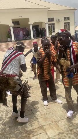 The video shows several men dancing in front of a building, possibly during a wedding celebration. There are at least six individuals present, dressed in a mix of traditional and modern clothing. One man in the foreground is wearing a white t-shirt with a striped sash, a fur-like garment around his waist, and objects attached to his legs that create a rattling sound when he moves. Other men are dressed in patterned shirts and trousers, with some wearing red hats or caps. Their movements suggest they are part of a coordinated dance or performance. The environment is a paved outdoor area, likely in front of a hall or event venue, as indicated by a banner visible in the background that reads "Anika & Nimnan". The building has a light-colored facade with multiple windows and doors, and there are other people in the background observing the performance. A truck is partially visible on the left side of the frame. The overall atmosphere appears festive and celebratory.