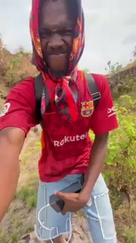 The video shows a man outdoors, standing on a large rock in a grassy area. The man has dark skin and is wearing a red Barcelona soccer jersey with the word "Rakuten" on the front, light-colored denim shorts with rips, and white sneakers. He also has a red and black patterned scarf covering his head and neck. He is carrying a black backpack. The man is holding the camera, and the view is from a low angle, looking up at him.  In the background, there is a rocky area with some green vegetation. The weather appears to be sunny. The man is looking directly at the camera. He then leans back to show the view.