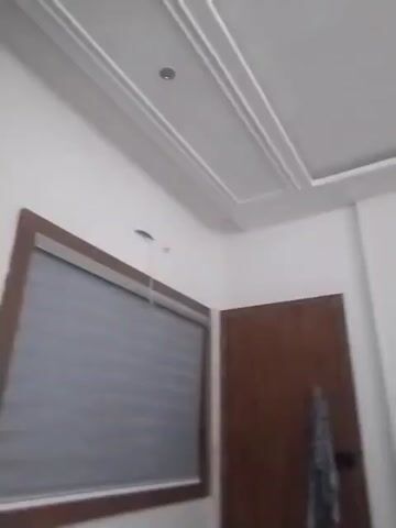 The video shows an interior of a room. The camera is pointed upwards towards the ceiling and then pans across the room. The ceiling has decorative molding. A white cylindrical object is hanging from the ceiling, possibly a light fixture or a camera. There is a wooden door on the left side of the frame. A gray, paneled wall is visible on the right side of the frame.