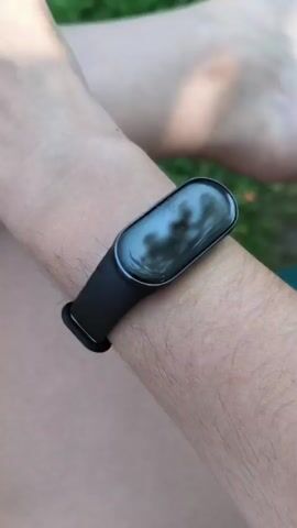 This is a close-up, vertical, eye-level shot of a person's wrist wearing a black fitness tracker. The fitness tracker displays the time as 4:20 PM, the date as Saturday, June 20th, a temperature of 28 degrees Celsius, a heart rate of 83, and icons for calories burned, steps taken, and battery percentage. The person's arm and a portion of their leg are visible, and the background consists of out-of-focus green grass and possibly some skin. The video is short, approximately 3 seconds long.