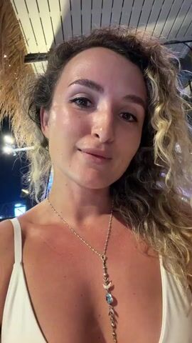 A woman with curly blonde hair and light skin is looking at the camera. She is wearing a white swimsuit and a silver necklace with a moon and celestial charms. Her hair is slightly messy, and she is smiling. The background appears to be an outdoor setting, possibly a restaurant or bar, with a thatched roof and lights. In the background, there is a fan and some greenery. The woman raises her right arm and waves.