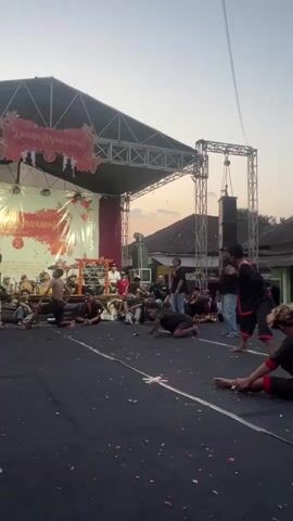 A group of people dressed in black and red are performing on a dark surface in front of a stage. The stage is decorated with a banner that says "Dharmayusawu" in red and orange. There are speakers and lighting equipment on the stage. Several people are sitting or kneeling on the dark surface, while others are standing. Some of the people on the ground appear to be moving, possibly in a dance or martial arts performance. In the background, there are buildings and trees, and the sky is a light orange, indicating it is either sunrise or sunset. The overall atmosphere appears to be a cultural performance or event.