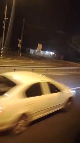 The video captures a nighttime view from a moving vehicle, likely a car, driving along a road. The scene is illuminated by streetlights and the headlights of passing vehicles.

**Subjects and Scene:** The primary focus is the road itself, with multiple lanes and a metal guardrail separating them. In the background, a low-rise building complex with several illuminated windows is visible, set back from the road. A signpost with text is also present near the building, though the text is not clearly legible. Trees and utility poles are scattered throughout the landscape, indicating an area that is not entirely urban. A white vehicle is partially visible in the right lane, moving in the same direction.

**Activity:** The activity is a journey by vehicle, as evidenced by the perspective and motion blur. The car is traveling at a moderate speed.

**Notable Details:**
*   **Time of Day:** Nighttime, with dark skies and artificial lighting.
*   **Weather:** Not explicitly discernible, but the lighting suggests clear conditions.
*   **Location Cues:** The prompt provides the location as Zavidovo, Russia. The building style and road infrastructure are consistent with a location in Russia.
*   **Emotions:** The video conveys a sense of travel and movement, with a slightly muted and cool color palette associated with nighttime.

**Visible Text:** A signpost is visible in the background near the building complex. While the text on the sign is not legible enough to be read, it appears to be a rectangular sign with dark text on a light background.