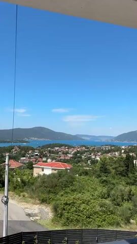 The video is a vertical shot of a coastal town with mountains in the background under a bright blue sky. In the foreground, there is lush green foliage and the roof of a building. As the video progresses, the camera pans up slightly to reveal an airplane flying in the distance. The airplane is white with a dark tail and appears to be in mid-flight. The water in the background is calm and blue, with a few boats visible. The overall scene suggests a clear, sunny day in a scenic location.