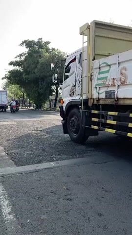 A motorcycle rider wearing a helmet and a vest is seen in the foreground, on the left side of the frame. The rider is on a scooter and appears to be stopped or moving slowly. In the background, a large truck is visible. The truck has a white cargo box with some writing on it, which appears to be faded. The truck also has yellow and black striped safety bars along its side. Further in the background, there are trees, buildings, and other vehicles, including another truck loaded with what appears to be sugarcane. The scene is outdoors, under a bright sky, suggesting it is daytime. The road surface is visible and appears to be asphalt.