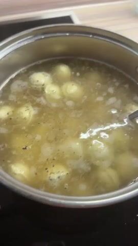 The video shows a close-up of a pot of food being cooked on a stovetop. The pot is filled with boiling water, in which many small, round dumplings are submerged. The dumplings are pale yellow in color and appear to be floating in a broth that has some specks of seasoning, possibly pepper, visible. A spoon is inserted into the pot and lifts some of the dumplings out of the water, showing them in detail. The stovetop appears to be an induction cooktop, with red indicator lights visible. The background is slightly blurred but suggests a kitchen environment with a countertop and possibly a backsplash. The overall scene depicts the process of cooking dumplings.