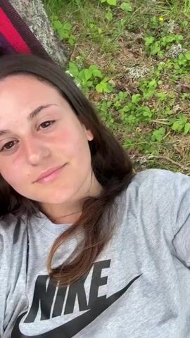 A young woman with dark hair, wearing a grey Nike t-shirt, lies in a striped hammock. She smiles and closes her eyes as the camera pans up to show tall trees and a blue sky with clouds. The setting appears to be outdoors, possibly a park or forest, with green foliage in the background. The video captures a peaceful moment of relaxation.