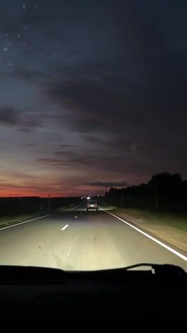 The video shows a first-person perspective of driving on a paved road at dusk. The sky is a deep blue with streaks of orange and pink from the setting sun. The headlights of the vehicle illuminate the road ahead, casting bright beams that blur the white lane markings. Another vehicle, a dark-colored car, is visible in the distance, its taillights glowing red. The road is flanked by dark trees and fields, with a few distant structures that could be utility poles or wind turbines. A dashboard display is partially visible in the lower part of the frame, showing some numerical data and indicators. The overall impression is one of a solitary drive through a twilight landscape.