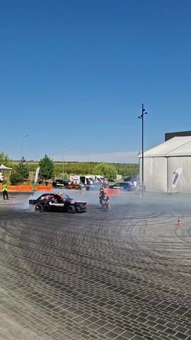 A black drift car with flames visible on its rear bumper is performing a donut maneuver on a paved area, creating a large cloud of white smoke. A person is standing on the rear of the car, waving their arms. Another person is on a motorcycle, also in the smoke cloud. Several other cars and people are visible in the background, as well as a large white tent and trees under a clear blue sky. Orange cones are placed on the ground around the area.